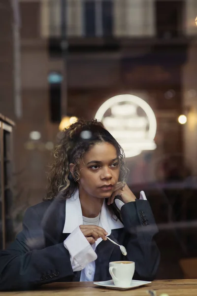 Stylish african american woman in wireless earphone listening music and holding spoon near cup of coffee in cafe — Stockfoto