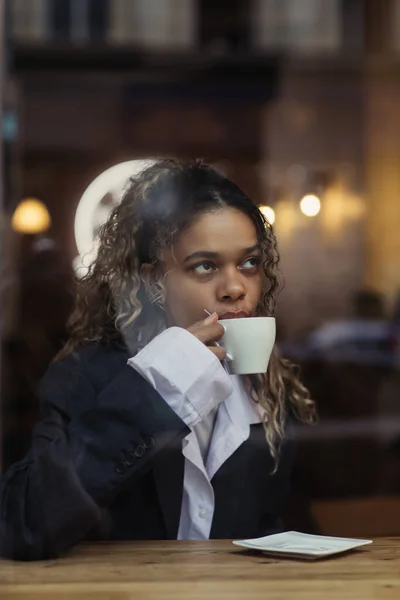 Stylish african american woman drinking coffee from cup behind window glass in cafe — Stockfoto