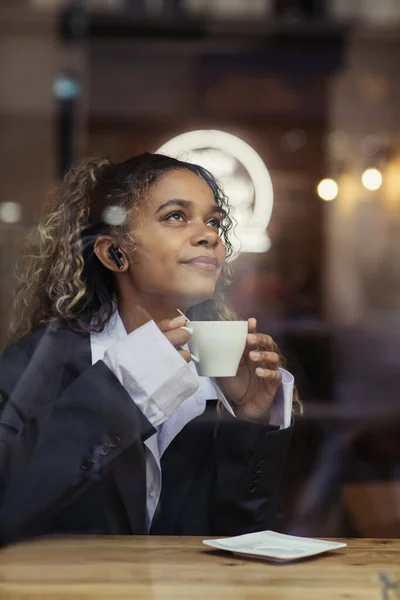 Dreamy african american woman in wireless earphone listening music and holding cup of coffee in cafe — Stock Photo