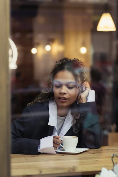 Curly african american woman holding cup of coffee behind window glass in cafe — Stock Photo