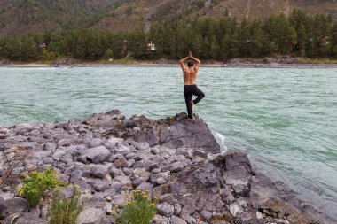 Nehir kıyısında yoga yapan bir adam, ağaç duruşu, dengede, tek ayak üzerinde duran.