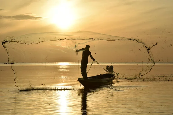 A fisherman is throwing a net from a fishing boat with gold twilight sky in background