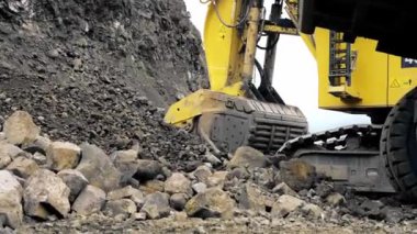 A large yellow caterpillar excavator is collecting soil and ore into the bucket. A quarry, mining of metals. Shooting from the ground. 4K
