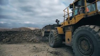 A yellow excavator with soil in a bucket drives away from a mound. Shot is made behind the excavator. A quarry, mining of metals. 4K