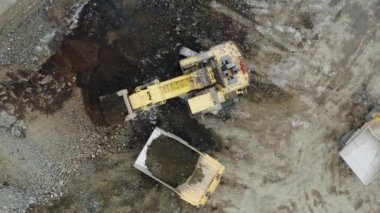 Top view of a yellow excavator that is loading ore into a dump truck. The drone flies off and makes a circular panoramic shot. A quarry. Mining industry. 4K