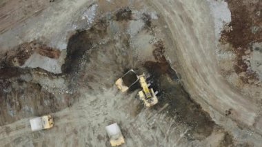 Yellow excavator in a quarry next to 3 heavy trucks. An excavator offloads ore into a dump truck. View from the drone, vertical panorama in motion. 4K