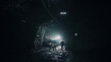 Three mining workers, in helmets, go underground through a dark tunnel. Two men and a woman. Flashlights on the helmets are on. 4K