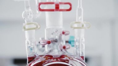 Close-up of boiling red liquid in a round glass flask. Production of food colorings. Bright laboratory. Chemical reactions. Vertical panorama. Camera moves from top to bottom. 4K