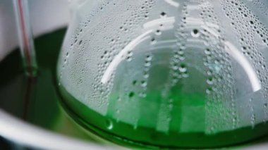 Misted glass flask with green liquid. Close-up. Drops flow down the flask. Chemical reaction. Food colorings production. Laboratory. Horizontal panorama. Camera moves from left to right. 4K