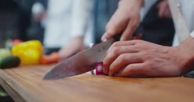 A cook in a white suit is slowly slicing a red onion on a wooden board. Beautiful dark kitchen. Close-up of hands and knife. Other chefs are cooking in the background. Professional cooking. 4K