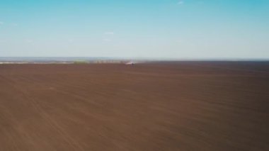 Kahverengi tarla üzerinde helikopter uçuşu. Kırmızı harrow 'lu mavi bir traktör tohum ekmek için toprağı sürüyor. Traktörün arkasında büyük bir toz tabakası uçar. Açık mavi gökyüzü. Güneşli bir gün. Yeşil alanlar ve uzaktaki sık ağaçlar. 4K