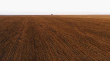 Kahverengi tarla üzerinde helikopter uçuşu. Kırmızı harrow 'lu mavi bir traktör tohum ekmek için toprağı sürüyor. Traktörün arkasında büyük bir toz tabakası uçar. 4K