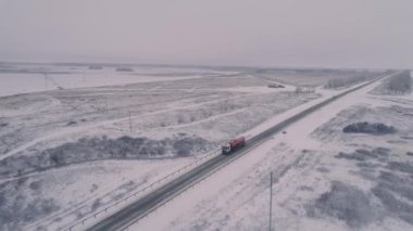 Karla kaplı kış otoyolunun en üst manzarası. Kırmızı tankeri olan bir kamyon şoförü. Uzun çıplak ağaçlar. Kar tarlasında elektrik hatları. Misty Sky. Helikopterden görüntü. 4K