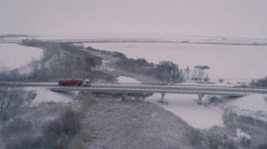 Köprüden geçen kırmızı tankerli bir tanker. Kış yolu. Elektrik hatları. Sütunlar. Kar tarlası. Çıplak ağaçlar ve karla kaplı çalılar. İnsansız hava aracının yan görüntüsü. 4K