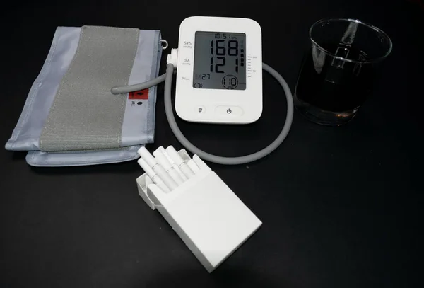blood pressure and heart rate monitor high blood pressure and heart rate measurements. cigarettes and alcoholic drink on the side. unhealthy lifestyle. black background