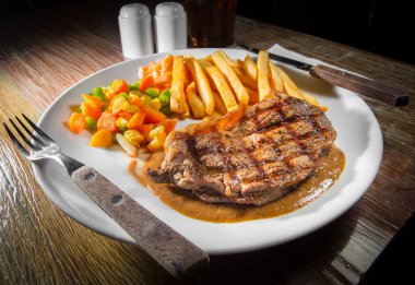Cooked grilled steak on a plate with vegetables and French fries on a wooden table