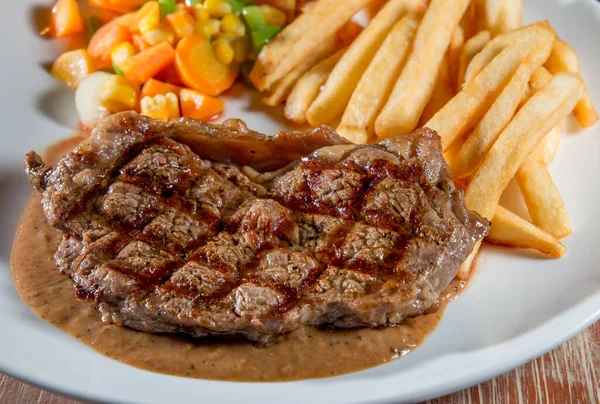Cooked grilled steak on a plate with vegetables and French fries on a wooden table