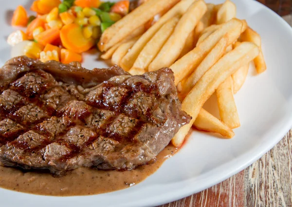 Cooked grilled steak on a plate with vegetables and French fries on a wooden table