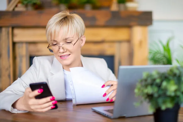 ノートパソコンのモニターの前に眼鏡をかけた家の中の女性が電話のメッセージに気を取られていた。日常生活における現代技術の概念. — ストック写真