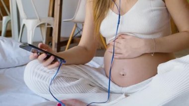 Genç hamile kadın kulaklıklı akıllı telefonuyla yatakta oturuyor. Çevrimiçi iletişim kuruyor. Akıllı telefonuyla yazıp mesaj gönderiyor..