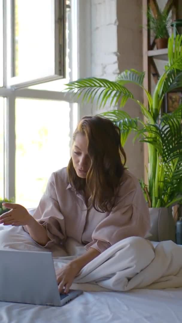 Vertikal video av en kvinna framför en laptop i sängen. Frilans - hon talar över videokommunikation och aktivt gester. — Stockvideo