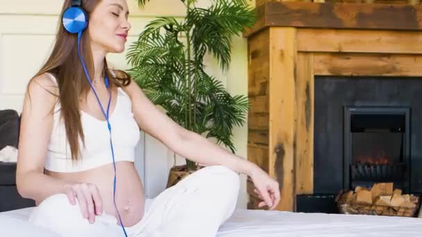 Беременная женщина слушает успокаивающую музыку через наушники. Она сидит дома на кровати и гладит живот.. — стоковое видео