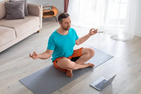Hombre tomando una clase de yoga en línea. Él está meditando sentado en el suelo delante de un monitor portátil. — Foto de Stock