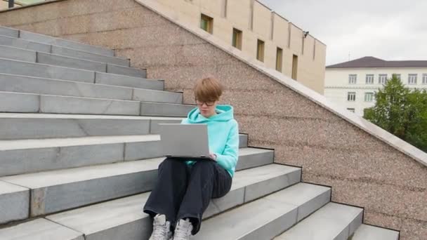 짧은 머리를 한 젊은 여자가 계단 위에 앉아 랩탑을 만지고 있습니다. 온라인 교육 개념. — 비디오