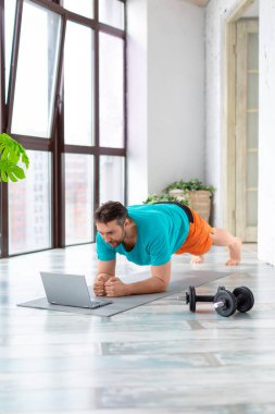 Orta yaşlı bir adamın dikey fotoğrafı. Laptop monitörünün önünde. Tahta egzersizi yapıyor. Online koçunun rehberliğini takip ediyor..