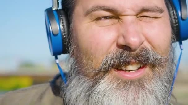 Homme barbu d'âge moyen avec écouteurs écoute de la musique, chante le long et hoche la tête au rythme de la musique. Gros plan sur le visage. — Video
