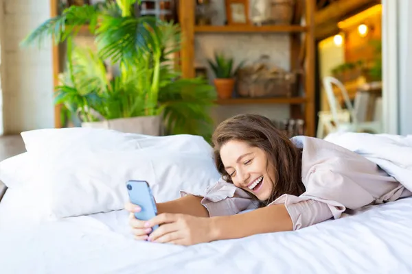 Foto seorang wanita tertawa bahagia dengan smartphone di tempat tidur. — Stok Foto