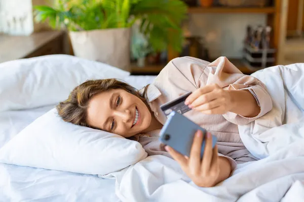 Wanita berbaring di tempat tidur membuat pesanan melalui aplikasi mobile. Dia menelepon nomor kartu untuk pembelian online. — Stok Foto