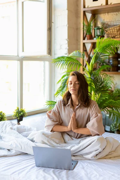 Vertikales Foto einer erwachsenen Frau, die im Bett vor dem Laptop-Monitor meditiert. Yoga-Praxis. — Stockfoto