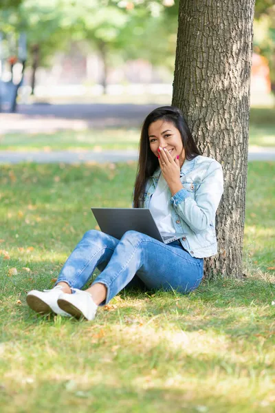 Pionowe zdjęcie młodej Azjatki siedzi w parku na trawie z laptopem. Wideo - śmieje się, zakrywając usta dłonią. — Zdjęcie stockowe