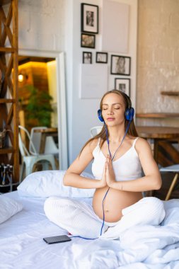 Genç hamile bir kadın mavi kulaklık takarak yatakta oturuyor ve online akıllı telefon uygulaması aracılığıyla meditasyon için müzik dinliyor..