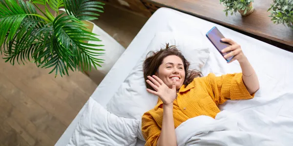 Cinta virtual (Virtual love): seorang wanita berbaring di tempat tidur dengan ponsel. Panggilan online untuk pacar virtual. Format foto 2x1. — Stok Foto