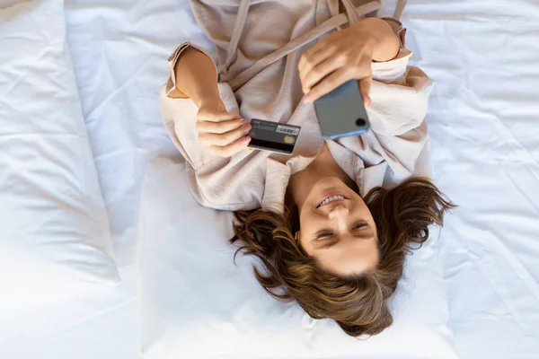 침대에 누워 있는 여자를 위에서 내려다보면 모바일 앱을 통해 주문을 할 수있다. 그녀는 온라인 구매를 위한 카드 번호를 검색 합니다.. — 스톡 사진