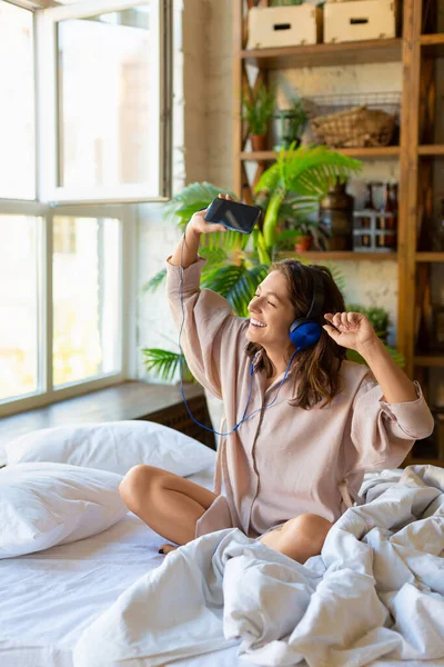 한 성인 여성이 일어나서 침대에 앉아 헤드폰으로 음악을 듣는다. — 스톡 사진