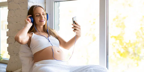 Femme enceinte dans un casque bleu assis sur le rebord de la fenêtre à la maison. Elle a un appel vidéo en ligne - consultation en ligne avec un médecin ou réunion en ligne avec son mari. — Photo