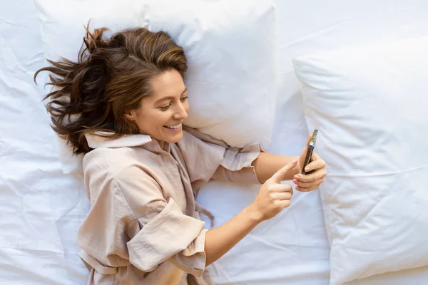 침대에 스마트폰을 가지고 있는 35 세 여성이 웃는 모습의 맨 위 사진. — 스톡 사진