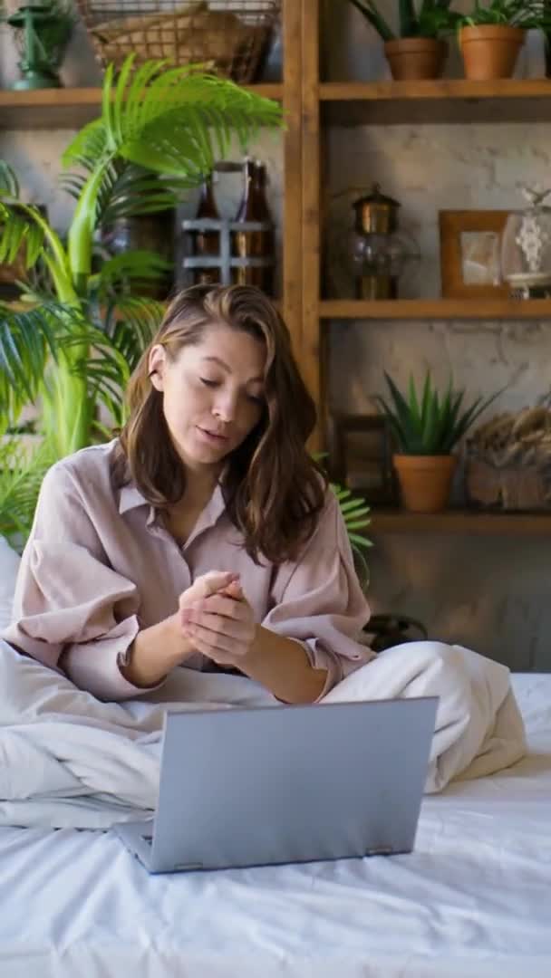 Vertikales Video einer Frau vor einem Laptop-Monitor im Bett. Freelance - sie spricht über Videokommunikation und aktive Gesten. — Stockvideo