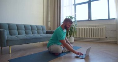 İnternette yoga dersi alan orta yaşlı bir adamın fotoğrafı. Dizüstü bilgisayar monitörünün önünde yerde oturmuş meditasyon yapıyor. 4K görüntü.