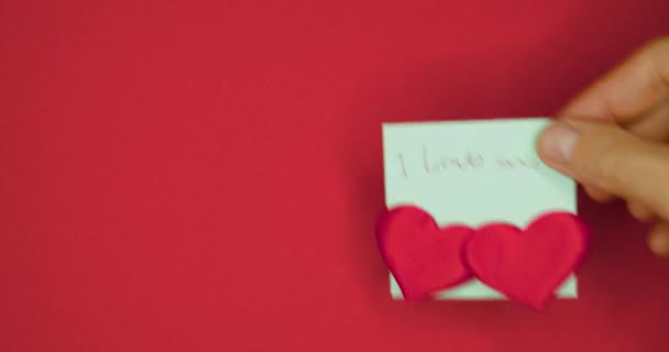 Mână de sex masculin lipește autocolant cu cuvintele pe care te iubesc scris în stilou și lipit două inimi pentru Valentine zi pe un fundal roșu. — Videoclip de stoc
