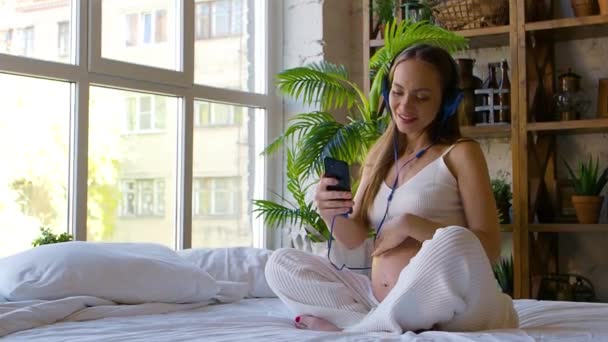 Een jonge zwangere vrouw zit op het bed met een smartphone in een koptelefoon en glimlacht. Ze communiceert actief online - schrijft en verstuurt berichten. — Stockvideo