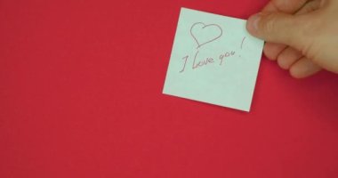Male hand is pasting sticker with the words I love you is written in pen and drawn with a heart for valentines day on a red background.