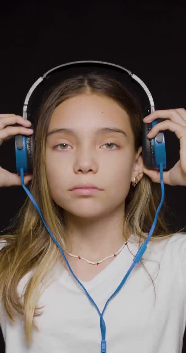 Teenage girl wearing headphones and listening to the hard rock music on a black background. Vertical video. — Video