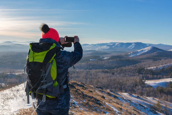 Hiker with backpack stands on the top of a mountain and takes pictures of the landscape with his smartphone. — Stockfoto