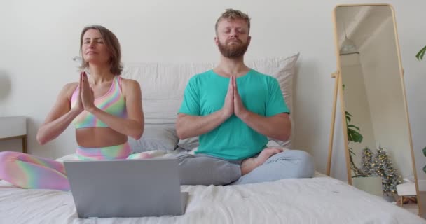 Middle-aged couple meditates in front of a laptop monitor in bed with guidance of an online mentor. — Stok video