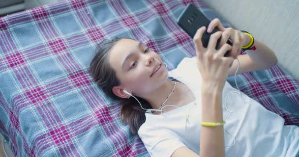 Vista superior de una adolescente se comunica con amigos en línea acostado en la cama con un teléfono inteligente. — Vídeos de Stock