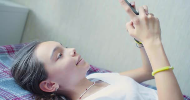 Teenagermädchen kommuniziert mit Freunden online auf dem Bett liegend mit einem Smartphone. — Stockvideo